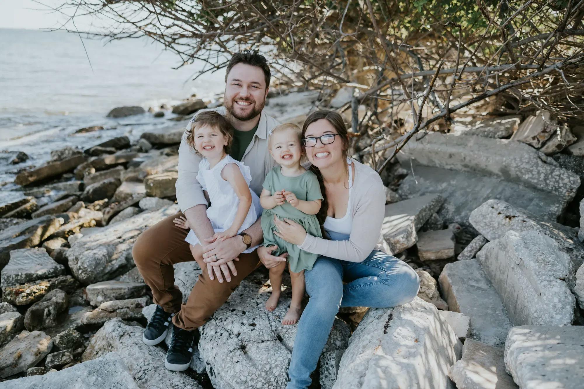 Photo of a dad, mom, and 2 young daughters sitting on rocks overlooking the water